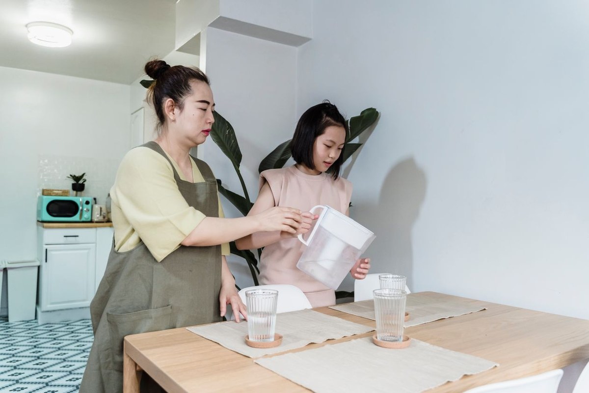 Mother and daughter pouring water from a white filter jug into glasses on a dining table