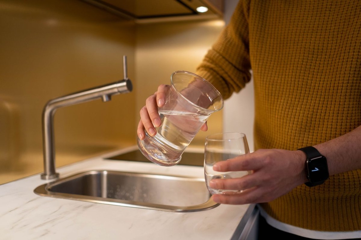 Person in a yellow sweater pouring clear water from a glass carafe into a tumbler at a kitchen sink